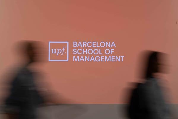 Two blurred figures walking past a UPF Barcelona School of Management projection.