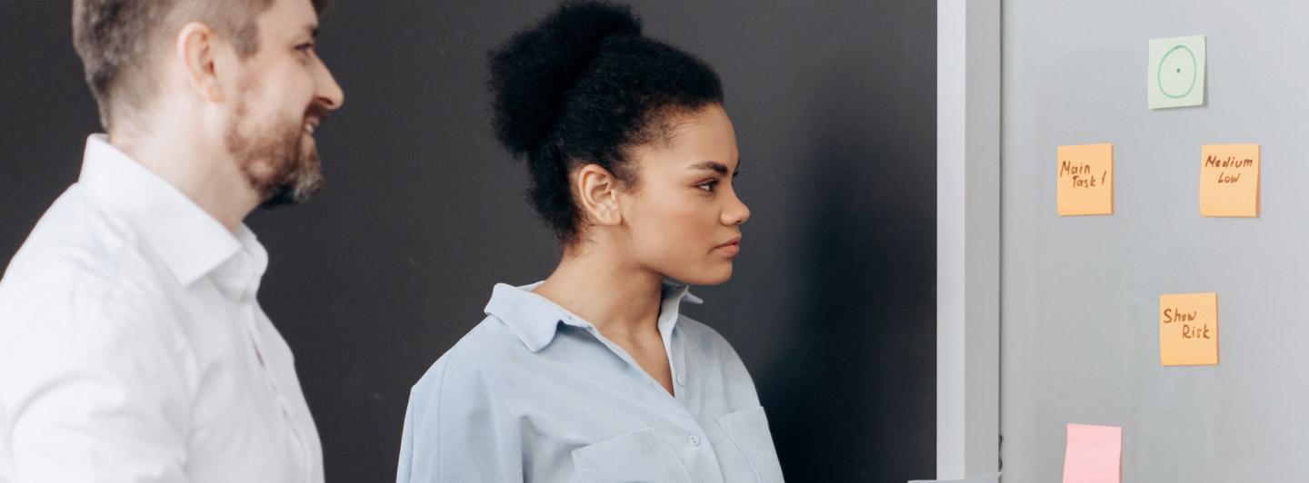 Two professionals discussing project tasks posted on a whiteboard with colorful sticky notes.