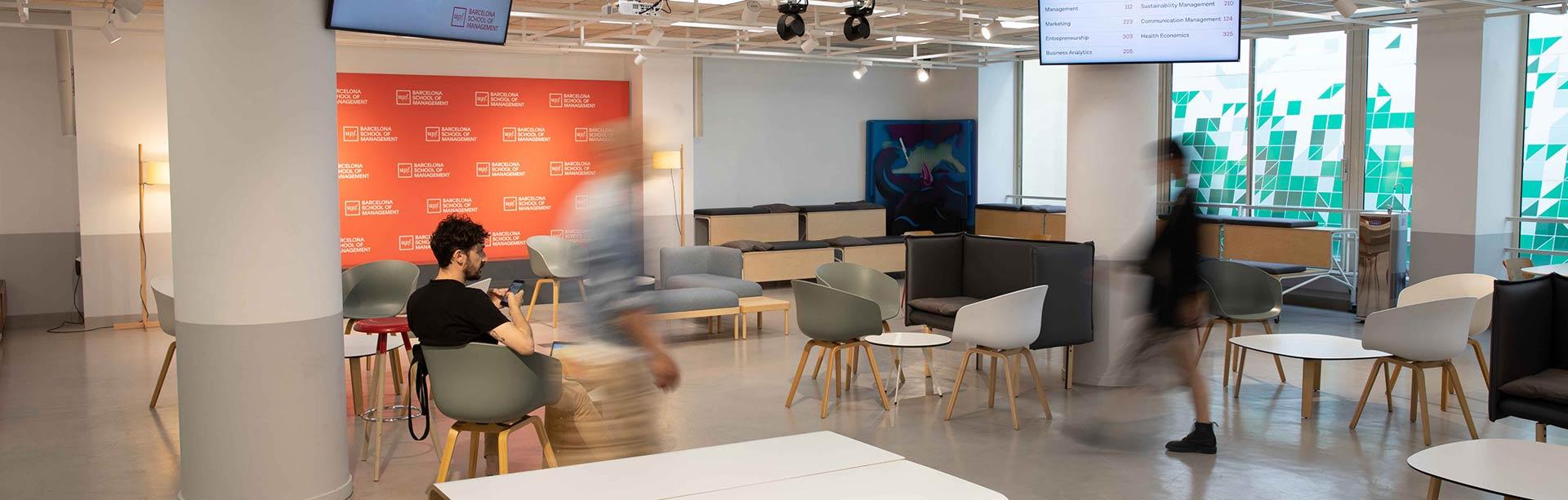 People walking through a modern university lobby with branded backdrop and seating area.